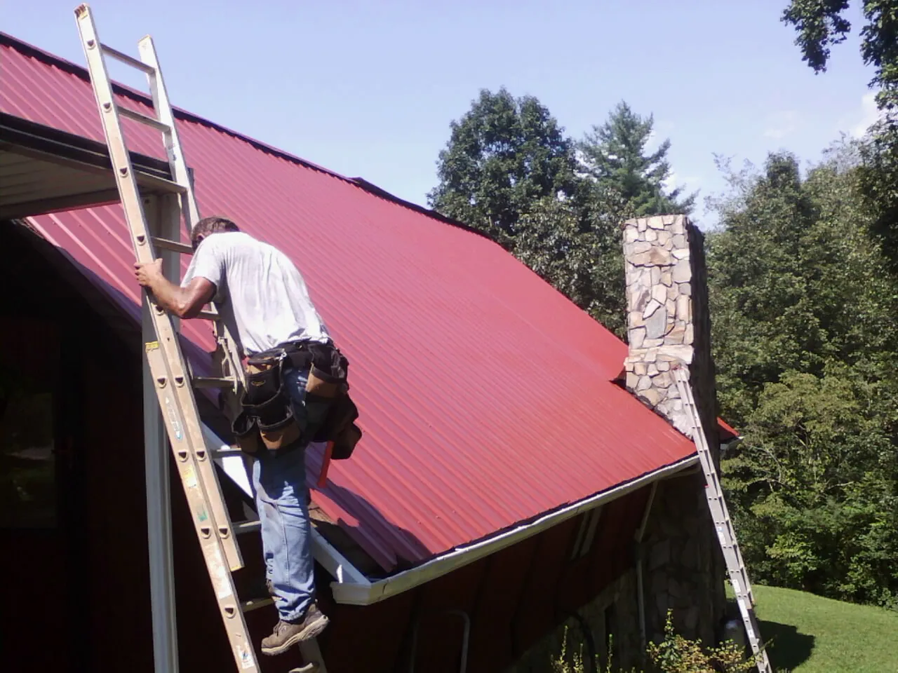 Roofer on a ladder beside a red metal roof and stone chimney — Soffit & Fascia Repair by Roofing Brothers in Hammett